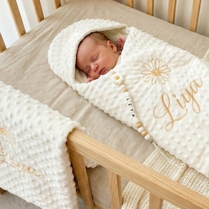 Könnte beinhalten: Ein Neugeborenes, eingewickelt in eine cremefarbene, strukturierte Decke mit einem Gänseblümchen-Design und dem Namen "Liya" in Gold gestickt. Das Baby liegt in einem Holzbettchen mit passender Decke.