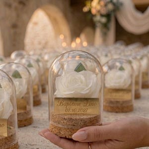 Può includere: Una rosa bianca racchiusa in una cupola di vetro, su una base di sughero. Un'etichetta dorata recita "Baptême Lino 01.10.2027". L'immagine mostra diverse bomboniere simili, suggerendo una celebrazione.