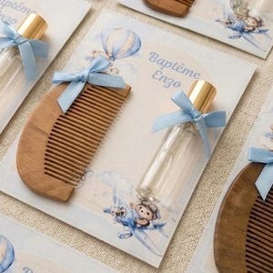 May include: A collection of baby shower favors featuring wooden combs and small perfume bottles. Each item is tied with a light blue ribbon and presented on a card with a watercolor illustration of a teddy bear in a hot air balloon. The card reads "Baptême Enzo".