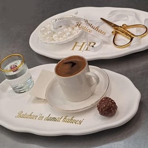 May include: Two white, oval-shaped serving trays with gold lettering and a small cup of coffee. One tray holds a small glass of water, a cup of coffee, and a chocolate truffle. The other tray has a ribbon, scissors, and pearls.