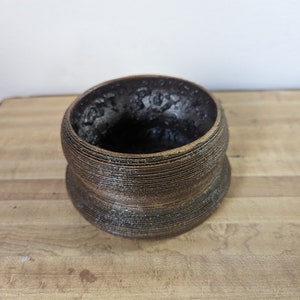 Puede incluir: Un macetero de cerámica marrón oscuro con un diseño texturizado y acanalado. El macetero tiene una abertura redonda ancha y una base ligeramente más estrecha.