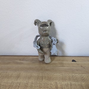 Puede incluir: Una figurita de cerámica de una criatura con un solo ojo, cuernos y una cadena en la mano. La figurita está pintada en tonos grises y marrones.