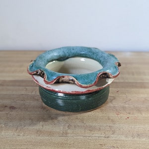 May include: A handmade ceramic bowl with a ruffled edge. The rim is a teal color, with a red accent. The bowl sits on a white base with a dark green bottom. The bowl is on a wooden surface.