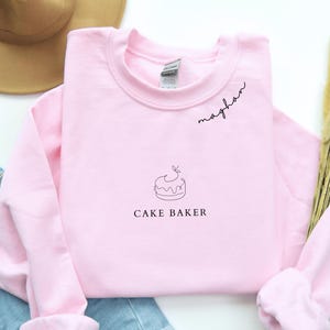 May include: A light pink sweatshirt with the word "meghan" in cursive script above a cake graphic and the words "CAKE BAKER" in a simple font. The sweatshirt is made of a soft material.