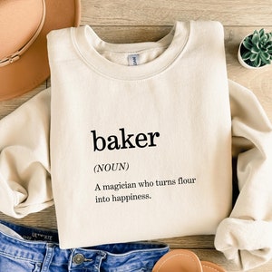 May include: A cream-colored sweatshirt with the text "baker (NOUN) A magician who turns flour into happiness." printed on the front.