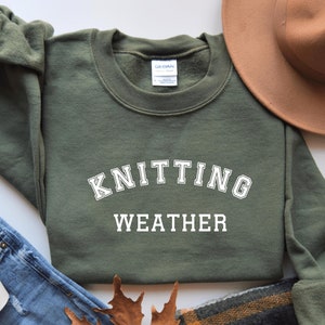 May include: A green sweatshirt with the text "Knitting Weather" in white letters on the front. The sweatshirt is folded and laid flat on a white surface.