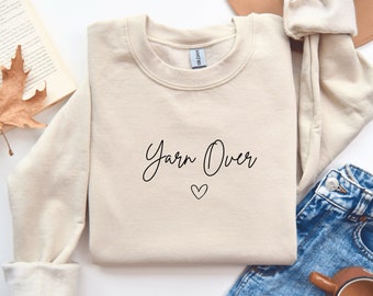 May include: A beige sweatshirt with the text "Yarn Over" in black script with a small heart below the text.