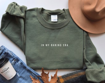 May include: A green sweatshirt with the text "IN MY BAKING ERA" in white lettering. The sweatshirt is laid out on a white surface with a brown hat, blue jeans, and a brown leaf.