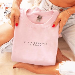 May include: A pink sweatshirt with the text "It's a good day to travel" in black lettering.