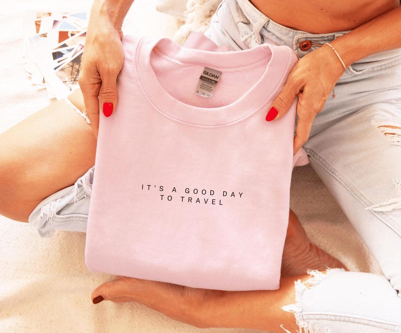 May include: A pink sweatshirt with the text "It's a good day to travel" in black lettering.