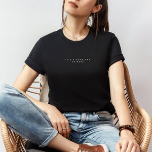 Pode incluir: Uma mulher vestindo uma camiseta preta com o texto "It's a good day to bake" em letras brancas. Ela est&aacute; sentada em uma cadeira de vime usando jeans azul.