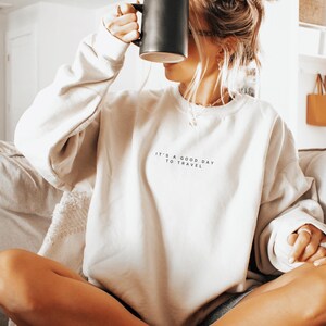 May include: A white sweatshirt with the text "It's a good day to travel" printed on the front. The person is wearing the sweatshirt and holding a black mug.