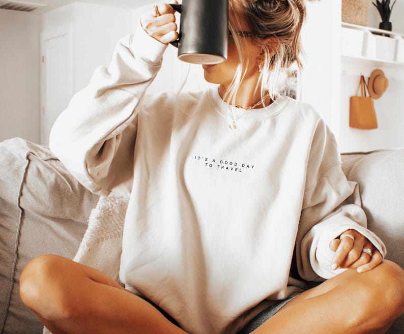 May include: A white sweatshirt with the text "It's a good day to travel" printed on the front. The person is wearing the sweatshirt and holding a black mug.