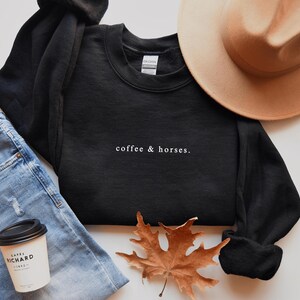 K&ouml;nnte beinhalten: Ein schwarzes Sweatshirt mit dem wei&szlig;en Text "coffee & horses". Das Bild zeigt auch blaue Jeans, einen beigen Hut, eine Kaffeetasse und ein braunes Herbstblatt.