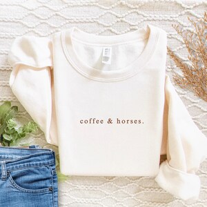K&ouml;nnte beinhalten: Cremefarbenes Sweatshirt mit dem Text "coffee & horses" in Braun. Das lang&auml;rmlige Shirt ist gefaltet, mit einer Jeans und dekorativen Pflanzen im Hintergrund. Das Shirt ist aus weichem Material.