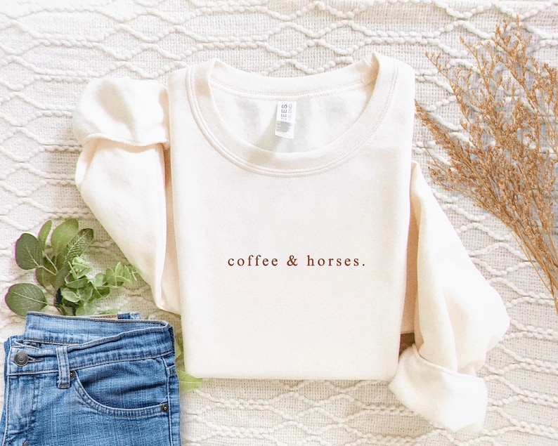 K&ouml;nnte beinhalten: Cremefarbenes Sweatshirt mit dem Text "coffee & horses" in Braun. Das lang&auml;rmlige Shirt ist gefaltet, mit einer Jeans und dekorativen Pflanzen im Hintergrund. Das Shirt ist aus weichem Material.