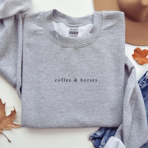 K&ouml;nnte beinhalten: Ein hellgraues Sweatshirt mit dem Text "coffee & horses." in schwarzer Schrift. Das Sweatshirt ist gefaltet, ein Teil des &Auml;rmels ist sichtbar. Ein brauner Hut, Jeans und ein Herbstblatt befinden sich im Hintergrund.