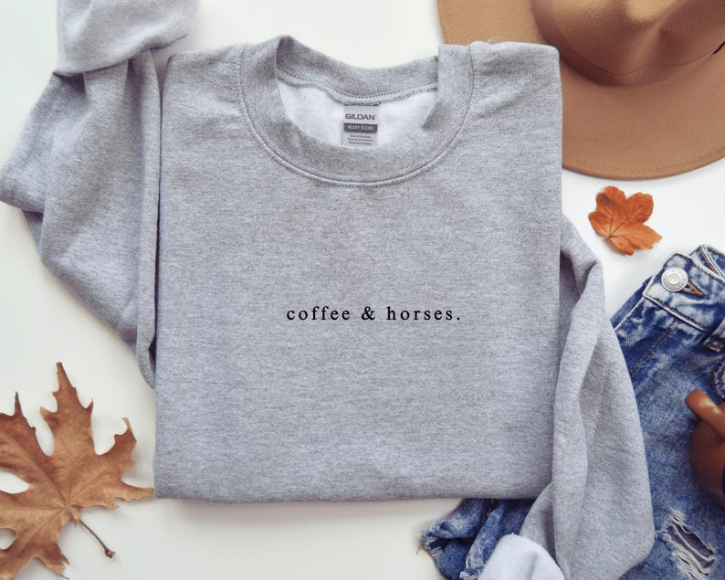 K&ouml;nnte beinhalten: Ein hellgraues Sweatshirt mit dem Text "coffee & horses." in schwarzer Schrift. Das Sweatshirt ist gefaltet, ein Teil des &Auml;rmels ist sichtbar. Ein brauner Hut, Jeans und ein Herbstblatt befinden sich im Hintergrund.