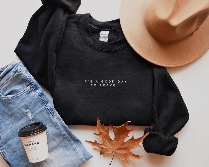 May include: Black sweatshirt with white text that reads "It's a good day to travel". The sweatshirt is laying on a white surface with a pair of blue jeans, a brown hat, and a fall leaf.