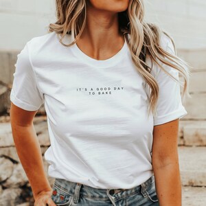 Pode incluir: Camiseta branca com o texto "It's a good day to bake" impresso na frente.