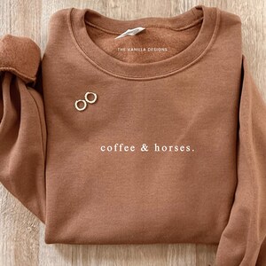 K&ouml;nnte beinhalten: Ein braunes Sweatshirt mit dem Text "coffee & horses." Das Sweatshirt hat einen Rundhalsausschnitt und lange &Auml;rmel. Zwei kleine goldene Creolen sind auf der Vorderseite platziert. Der Markenname "THE VANILLA DESIGNS" ist auf dem Etikett aufgedruckt.