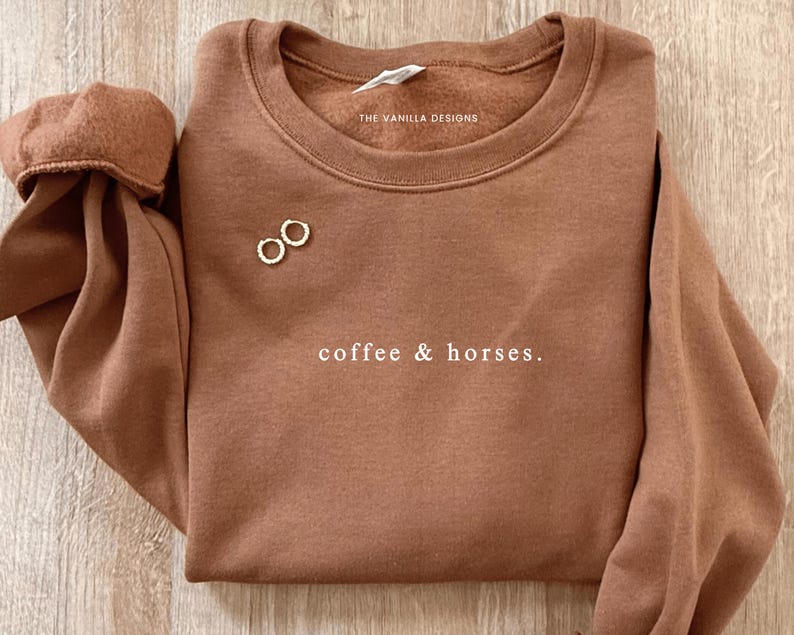 K&ouml;nnte beinhalten: Ein braunes Sweatshirt mit dem Text "coffee & horses." Das Sweatshirt hat einen Rundhalsausschnitt und lange &Auml;rmel. Zwei kleine goldene Creolen sind auf der Vorderseite platziert. Der Markenname "THE VANILLA DESIGNS" ist auf dem Etikett aufgedruckt.