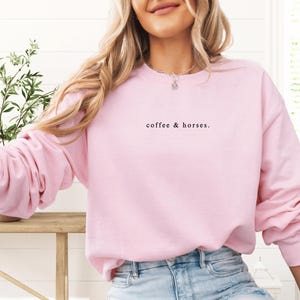 K&ouml;nnte beinhalten: Ein hellrosa Sweatshirt mit dem Text "coffee & horses." in schwarzer Schrift. Das lang&auml;rmlige Oberteil ist aus einem weichen Material gefertigt. Das Sweatshirt ist ein l&auml;ssiger Stil und wird von einer Person getragen.