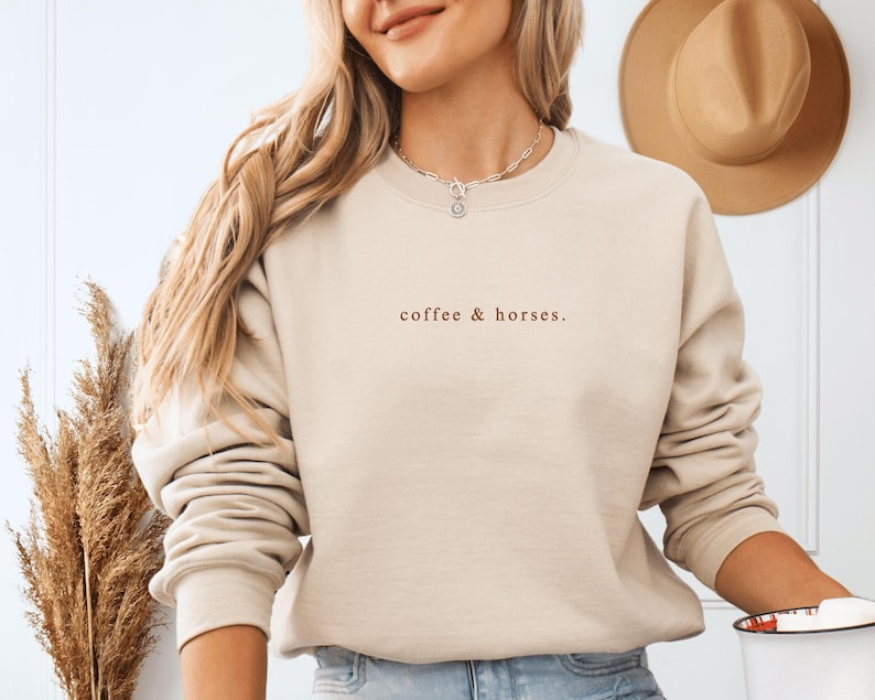 K&ouml;nnte beinhalten: Beiges Sweatshirt mit Rundhalsausschnitt und dem Text "coffee & horses" in Braun. Das Sweatshirt hat eine neutrale Farbe und lange &Auml;rmel. Im Hintergrund h&auml;ngt ein brauner Hut.