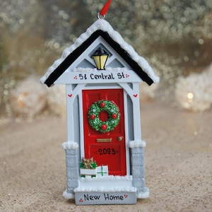 May include: A white and gray house-shaped Christmas ornament with a red door, a green wreath, and the text "51 Central St." and "New Home".