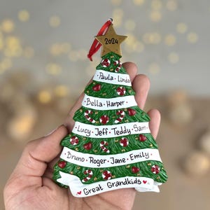 May include: A green Christmas tree ornament featuring white ribbons with the names Paula, Linda, Bella, Harper, Lucy, Jeff, Teddy, Sam, Bruno, Roger, Jane, Emily, and Great Grandkids. A gold star at the top is inscribed with the year 2024.