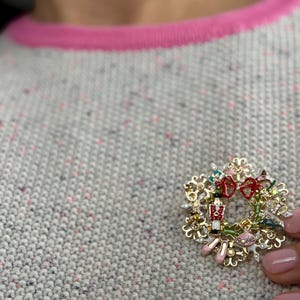 May include: A gold-toned Christmas wreath brooch with red bows, a toy soldier, and pink ballet slippers. The brooch is set against a gray sweater with a pink trim.