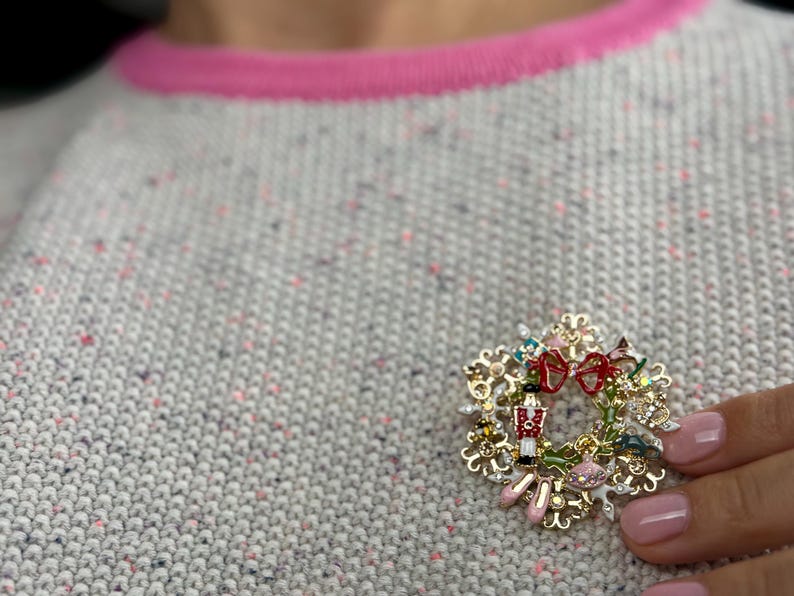 May include: A gold-toned Christmas wreath brooch with red bows, a toy soldier, and pink ballet slippers. The brooch is set against a gray sweater with a pink trim.