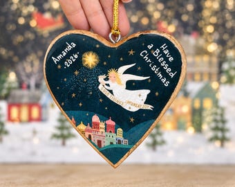 May include: A heart-shaped wooden ornament with a dark blue background featuring a golden star, an angel holding a banner that reads "GOOD TIDINGS OF GREAT JOY," and a small town. Text reads "Amanda .2026." and "Have a Blessed Christmas."
