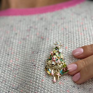 May include: A gold-toned Christmas tree brooch with colorful enamel details. The brooch features a star on top and is adorned with miniature ornaments. The tree is set against a gray sweater with pink trim.
