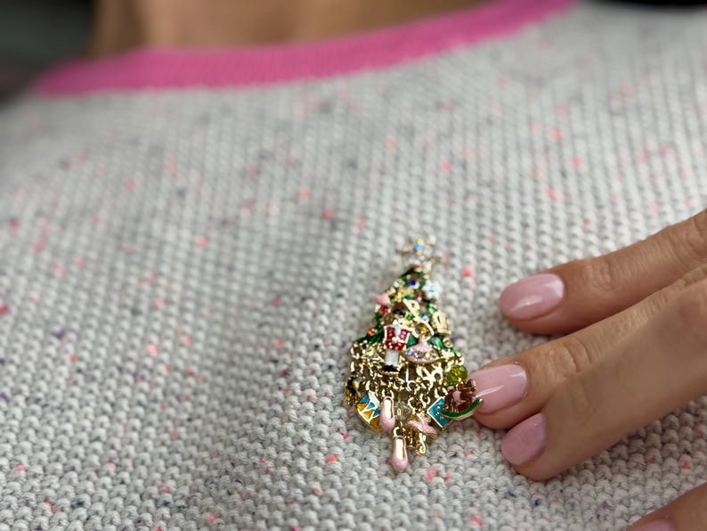May include: A gold-toned Christmas tree brooch with colorful enamel details. The brooch features a star on top and is adorned with miniature ornaments. The tree is set against a gray sweater with pink trim.