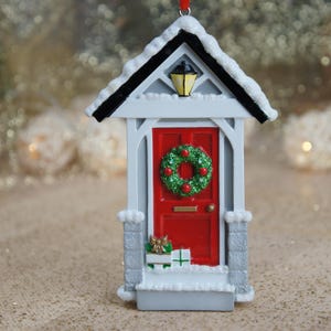 May include: White Christmas ornament shaped like a house with a red door, a green wreath with red berries, and a small porch with wrapped gifts.