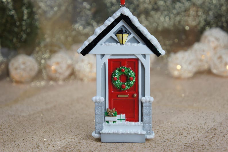 May include: White Christmas ornament shaped like a house with a red door, a green wreath with red berries, and a small porch with wrapped gifts.