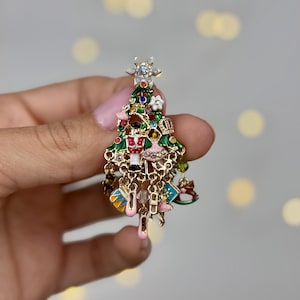 May include: A Christmas tree-shaped brooch with green, red, and gold enamel. The brooch features miniature ornaments, including a nutcracker, ballet slippers, and a crown. The tree is topped with a star and has dangling charms.