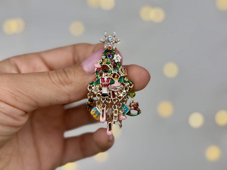 May include: A Christmas tree-shaped brooch with green, red, and gold enamel. The brooch features miniature ornaments, including a nutcracker, ballet slippers, and a crown. The tree is topped with a star and has dangling charms.