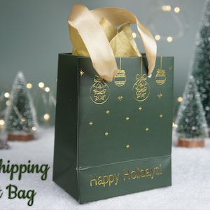 May include: A dark green gift bag with gold accents, featuring holiday ornament designs and the text "Happy Holidays!". The bag is filled with gold tissue paper and has a gold ribbon handle. Decorative Christmas trees and lights are in the background.
