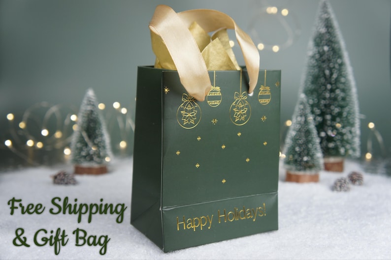 May include: A dark green gift bag with gold accents, featuring holiday ornament designs and the text "Happy Holidays!". The bag is filled with gold tissue paper and has a gold ribbon handle. Decorative Christmas trees and lights are in the background.
