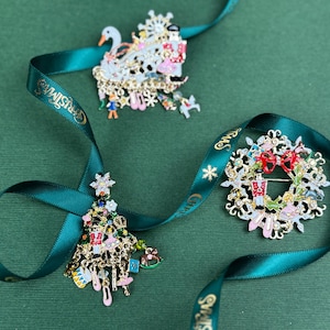 May include: Three Christmas-themed brooches with intricate details, including a swan, a wreath, and a Christmas tree. Each brooch is adorned with colorful accents and dangling charms. A green ribbon with gold text and stars weaves around the brooches.