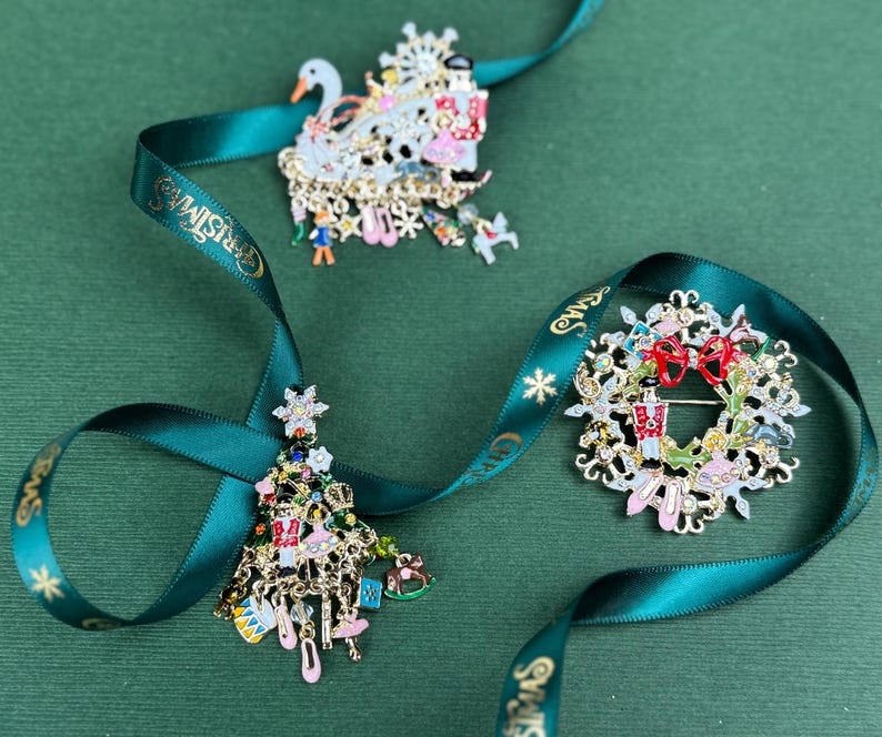 May include: Three Christmas-themed brooches with intricate details, including a swan, a wreath, and a Christmas tree. Each brooch is adorned with colorful accents and dangling charms. A green ribbon with gold text and stars weaves around the brooches.