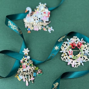 May include: Three Christmas-themed brooches: a swan, a wreath, and a tree, each with intricate details and colorful accents. A teal ribbon with "Christmas" text weaves through the brooches, set against a green background.