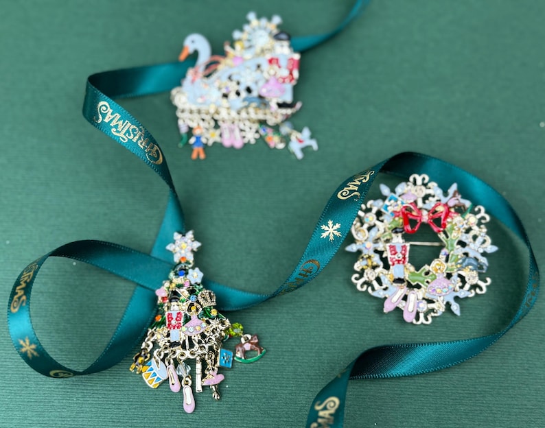 May include: Three Christmas-themed brooches: a swan, a wreath, and a tree, each with intricate details and colorful accents. A teal ribbon with "Christmas" text weaves through the brooches, set against a green background.