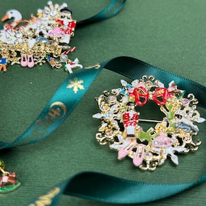 May include: Close-up of three Christmas-themed brooches. One features a swan, another a wreath with a nutcracker, and the third a Christmas tree. All are gold-toned with colorful enamel details and rhinestones, resting on a green surface with a green ribbon.
