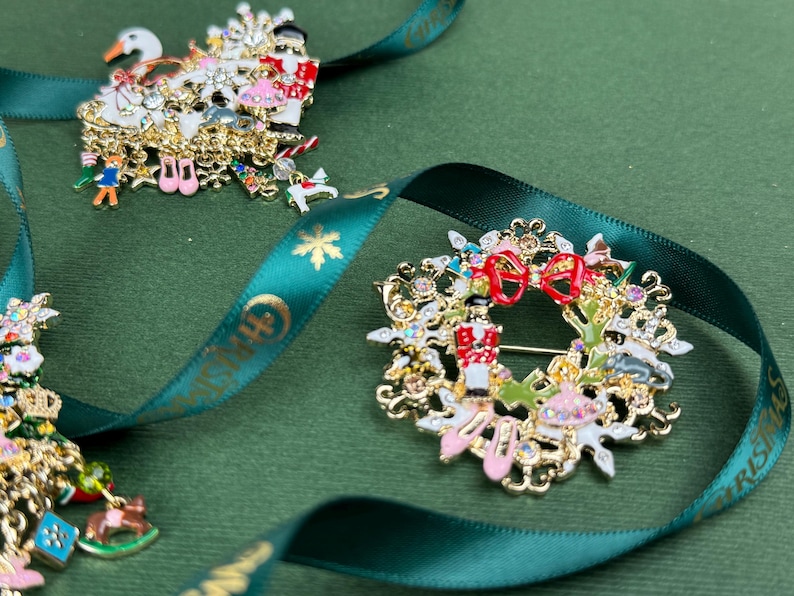 May include: Close-up of three Christmas-themed brooches. One features a swan, another a wreath with a nutcracker, and the third a Christmas tree. All are gold-toned with colorful enamel details and rhinestones, resting on a green surface with a green ribbon.