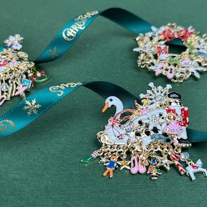 May include: Three Christmas-themed brooches with gold-tone metal and colorful enamel details. One is a Christmas tree, another a wreath, and the third depicts a swan and figures. All are on a green background with a teal ribbon that says "Christmas".