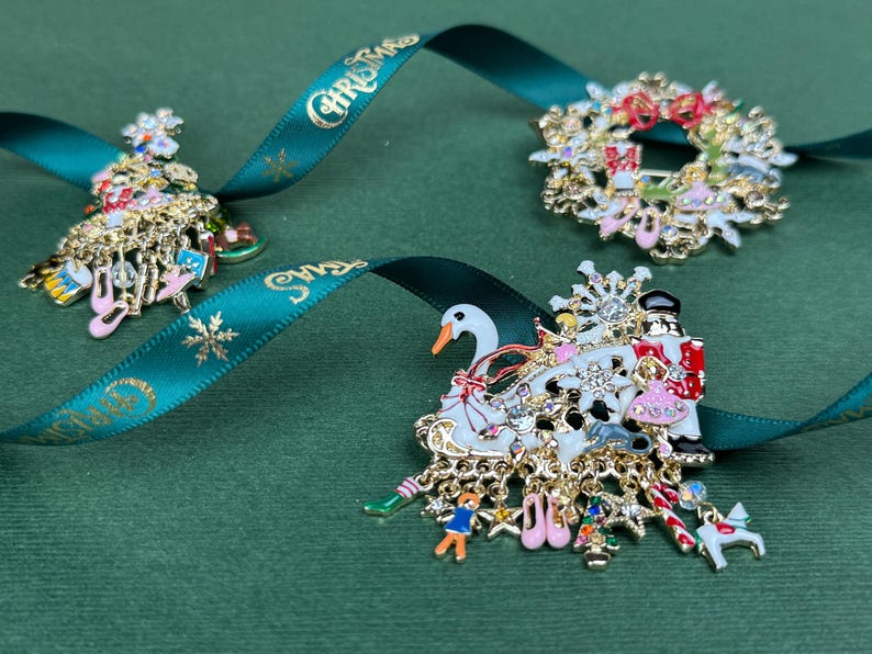 May include: Three Christmas-themed brooches with gold-tone metal and colorful enamel details. One is a Christmas tree, another a wreath, and the third depicts a swan and figures. All are on a green background with a teal ribbon that says "Christmas".