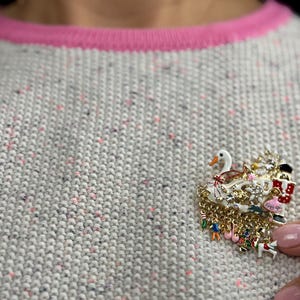 May include: A decorative brooch featuring a swan, Christmas tree, and other festive elements. The brooch is made of gold-toned metal with colorful enamel details and dangling charms. It is pinned to a gray sweater with a pink neckline.